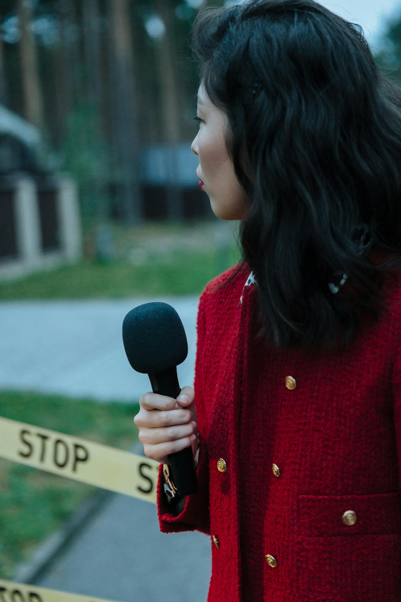 A female reporter in a red coat holding a microphone near a 'Stop' sign outdoors.