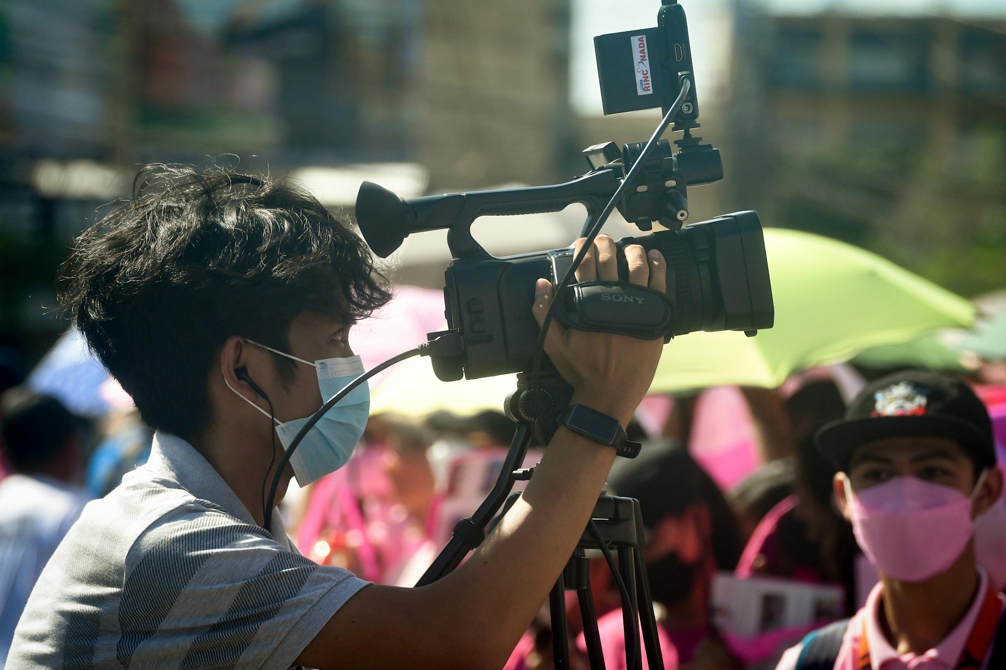 Cameraman wearing a mask films at a lively outdoor event, capturing live scenes.